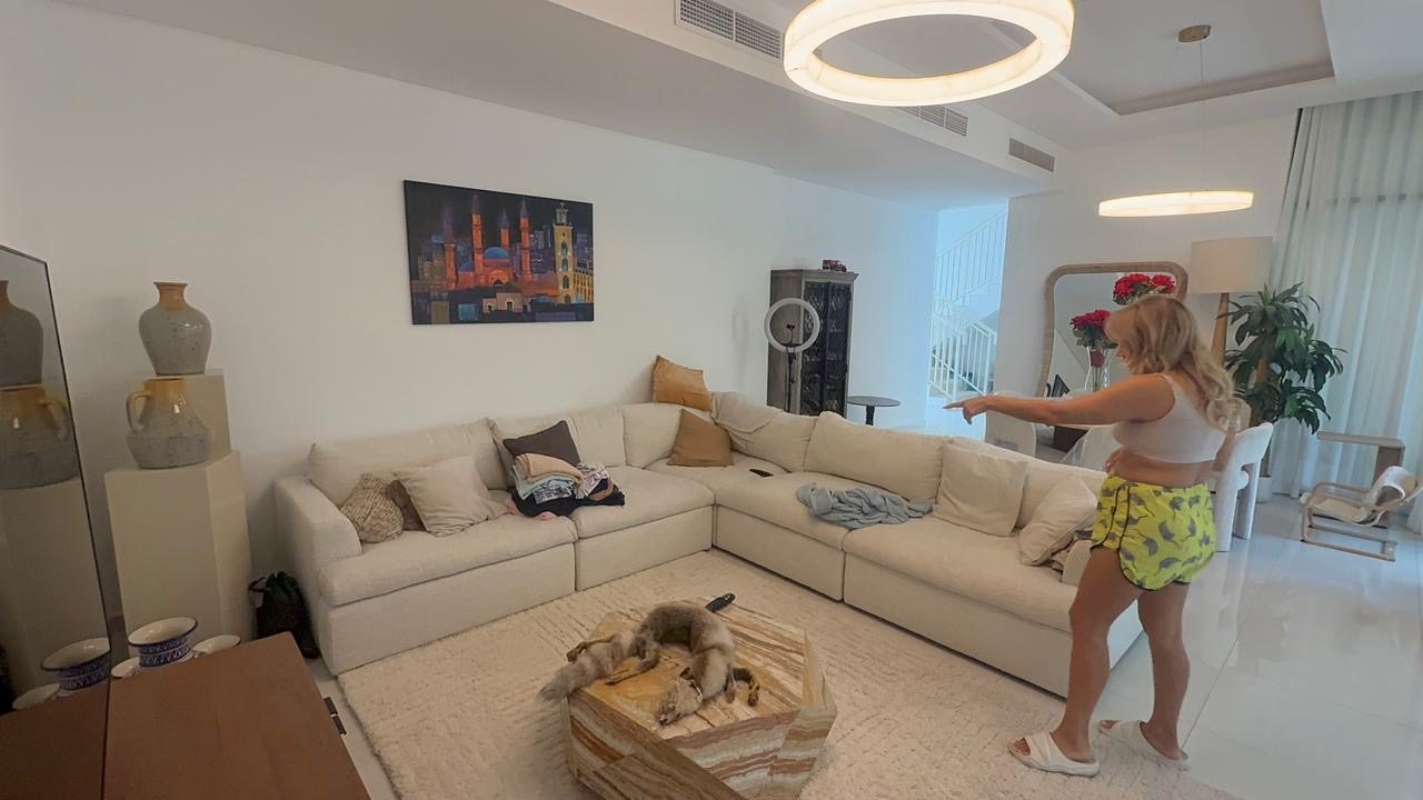 Villa living room as filmed on a phone, with a person standing in the room and clutter on the sofa